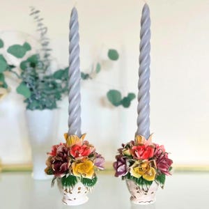 May include: Two gray, twisted taper candles sit atop floral ceramic candle holders. The candle holders are white with colorful flowers in shades of red, yellow, and purple. The candles are approximately 10 inches tall.
