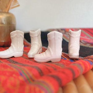 May include: Four small white ceramic cowboy boot shaped vases. The vases are sitting on a colorful woven rug.