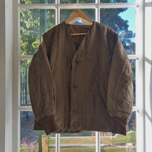 May include: A brown quilted jacket with a V-neck and button closure, hanging on a wooden hanger. The jacket has brown ribbed cuffs and is displayed in front of a window with a view of trees and blue sky.