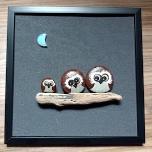 Peut inclure: Une œuvre d'art encadrée représentant trois figurines d'hibou peintes perchées sur un morceau de bois flotté. Les hiboux sont bruns avec des accents blancs et des yeux noirs. Un croissant de lune bleu se trouve dans le coin supérieur gauche de l'œuvre d'art.