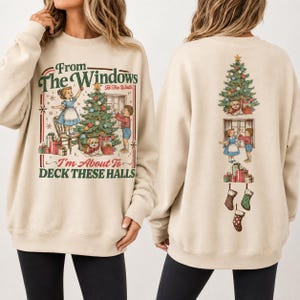 Puede incluir: Una sudadera beige de cuello redondo con un gráfico navideño de estilo vintage. El frente muestra niños decorando un árbol con el texto "From The Windows I'm About To DECK THESE HALLS". La parte posterior muestra iconos navideños apilados.