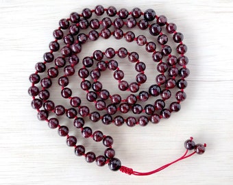 May include: A red garnet mala bead necklace with a red string tassel. The beads are round and have a natural, polished finish.