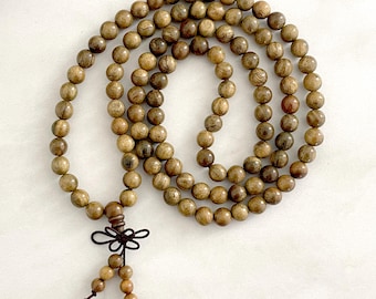 May include: A long strand of round, light brown wooden beads with a natural grain pattern. The beads are arranged in a circular loop and feature a dark brown decorative knot at the bottom, which connects to two smaller dangling bead accents.