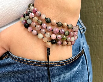 May include: Multi-colored gemstone mala bracelet with black beads and a black cord. The bracelet is wrapped around a person's wrist.