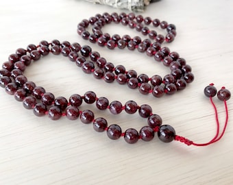 May include: A red gemstone mala necklace with a tassel. The necklace is made of small, round beads and is strung on a red cord.