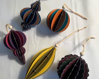 May include: A collection of five colorful, paper Christmas ornaments. The ornaments are various shapes, including a sphere, teardrop, and a decorative shape. Each ornament has a string for hanging and a wooden bead.