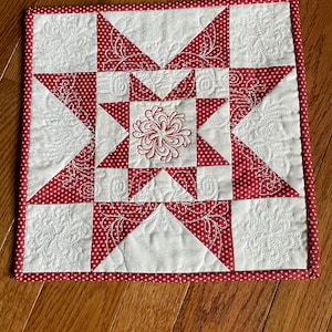 May include: A red and white quilted square with a star design in the center. The star is made up of white triangles with red polka dots and white triangles with a white floral design. The square is surrounded by a red border.