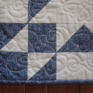 Puede incluir: Primer plano de una colcha con un patr&oacute;n azul y blanco. La colcha est&aacute; hecha de cuadrados y tri&aacute;ngulos, y la tela tiene un peque&ntilde;o estampado floral. La colcha est&aacute; acolchada con un patr&oacute;n de espiral.