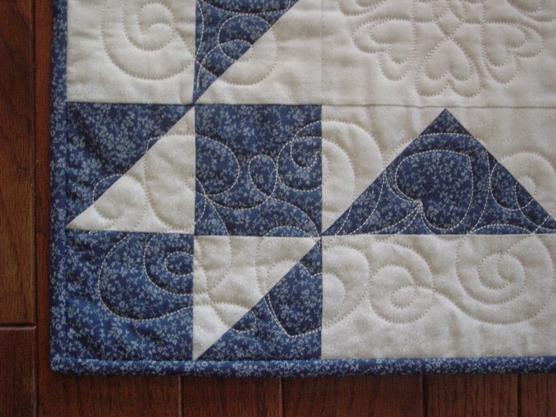Puede incluir: Primer plano de una colcha con un patr&oacute;n azul y blanco. La colcha est&aacute; hecha de cuadrados y tri&aacute;ngulos, y la tela tiene un peque&ntilde;o estampado floral. La colcha est&aacute; acolchada con un patr&oacute;n de espiral.