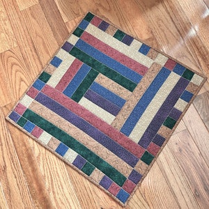 May include: A square patchwork rug with a geometric pattern in shades of green, blue, purple, and brown. The rug is made of different fabrics with a variety of textures.
