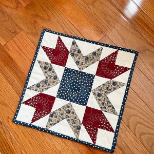May include: A red, white, and blue patchwork quilt square with a star design. The quilt is made of different fabrics with floral patterns and is quilted with a white thread.