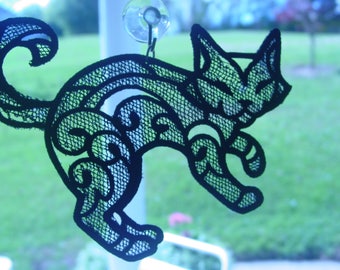 May include: Black lace cat silhouette hanging from a suction cup on a window. The cat is in a playful pose with its tail curled up.