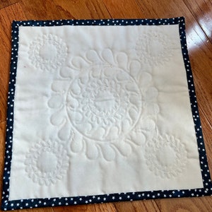 May include: A white quilted square placemat with a navy blue border featuring white stars. The placemat has a decorative quilted design of four circles with swirls.