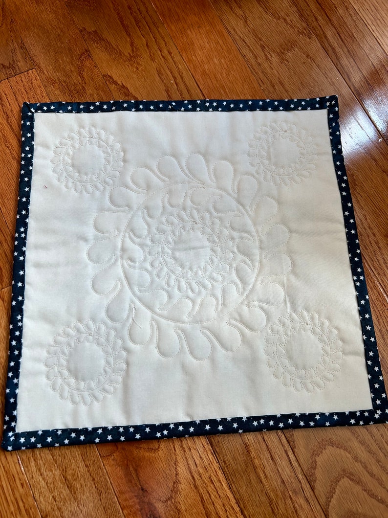 May include: A white quilted square placemat with a navy blue border featuring white stars. The placemat has a decorative quilted design of four circles with swirls.