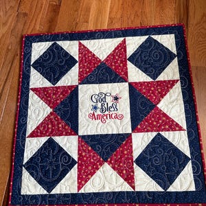 May include: A patriotic square quilt with a red, white, and blue star pattern. The quilt features a central embroidered text that reads "God Bless America". The design includes alternating star and diamond shapes.