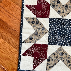 May include: A red, white, and blue patchwork quilt square with a star design. The quilt is made of different fabrics with floral and star patterns. The quilt is edged with a navy blue trim with white stars.