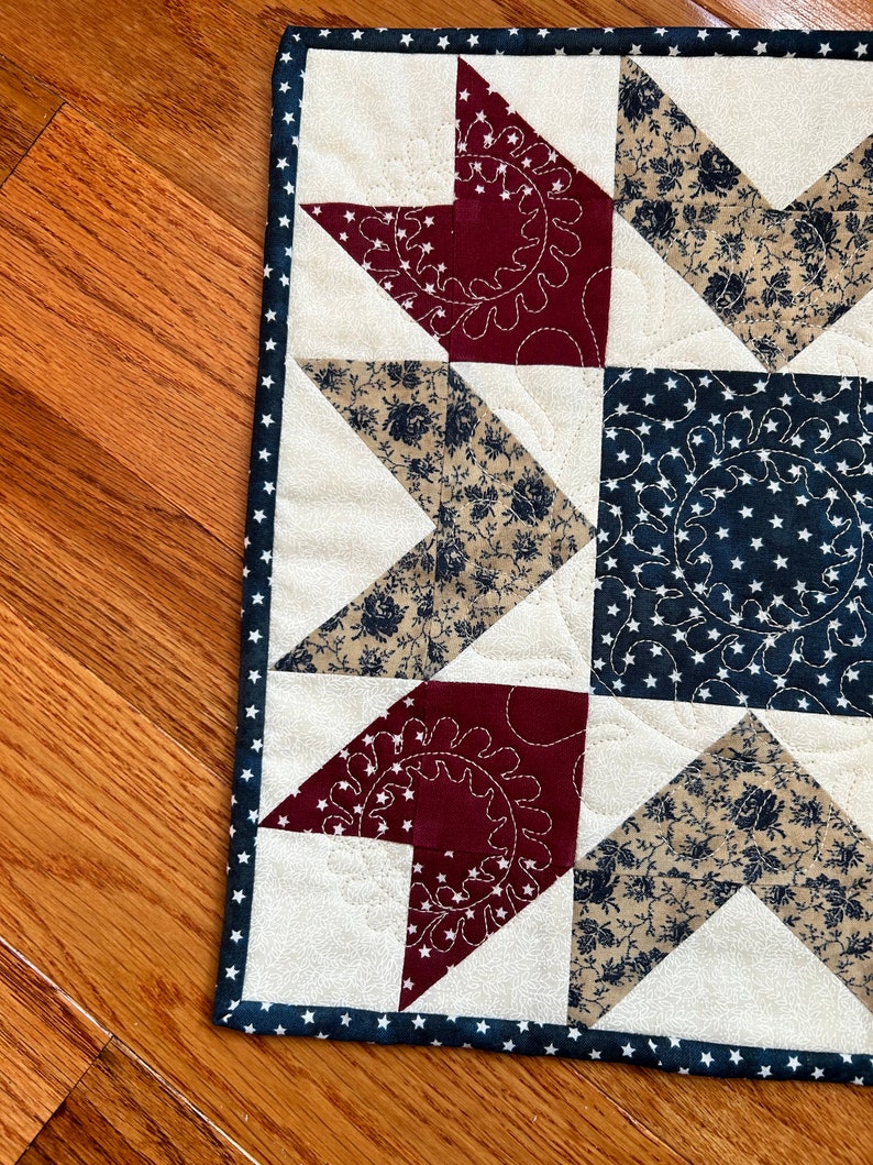 May include: A red, white, and blue patchwork quilt square with a star design. The quilt is made of different fabrics with floral and star patterns. The quilt is edged with a navy blue trim with white stars.