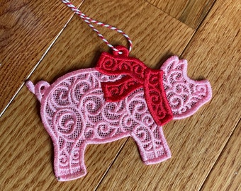 Puede incluir: Adorno en forma de cerdo rosa con bufanda roja, hecho de encaje bordado. El adorno tiene un patrón de remolino decorativo y una cuerda roja y blanca para colgar. El cerdo está sobre una superficie de madera.