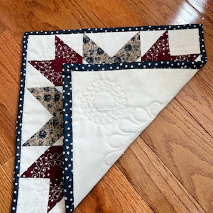 May include: A white quilted table runner with a navy blue and white star patterned border. The center of the runner is decorated with a quilted floral design. The runner is folded in half and lying on a wooden floor.