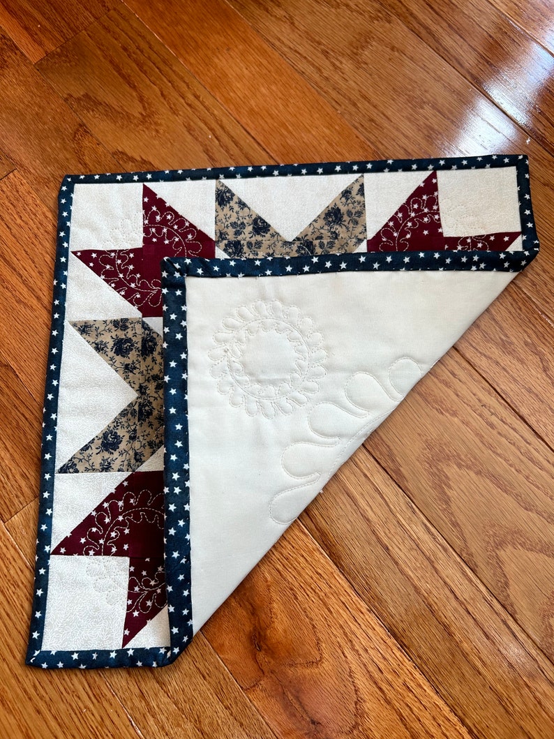 May include: A white quilted table runner with a navy blue and white star patterned border. The center of the runner is decorated with a quilted floral design. The runner is folded in half and lying on a wooden floor.