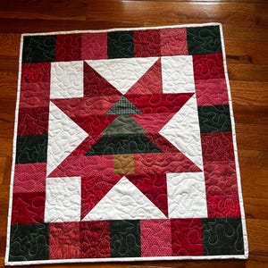 May include: A red, green, and white patchwork quilt with a star design in the center. The star is made up of smaller squares and triangles, and it features a green Christmas tree in the center. The quilt is edged with a white border.