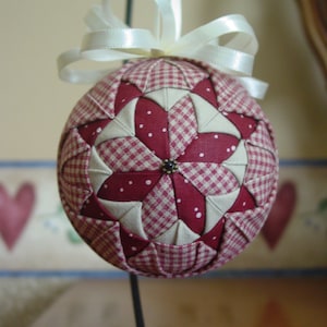 May include: A red and white gingham fabric Christmas ornament with a white satin ribbon. The ornament is shaped like a ball and has a starburst pattern.
