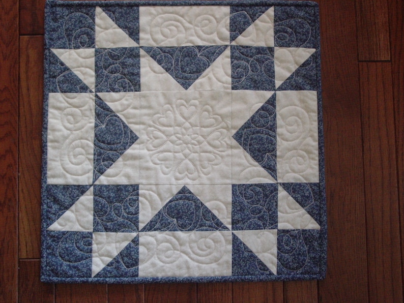 Puede incluir: Una colcha blanca y azul con un dise&ntilde;o de estrella en el centro. La colcha est&aacute; hecha de tela con un peque&ntilde;o estampado floral. La colcha est&aacute; acolchada con un dise&ntilde;o de remolino.