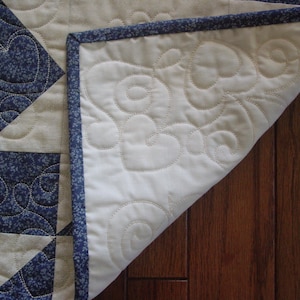 Puede incluir: Una colcha blanca con un patr&oacute;n floral azul y blanco. La colcha est&aacute; doblada por la mitad y se ve el lado de atr&aacute;s. El lado de atr&aacute;s es blanco con un borde azul y un patr&oacute;n de espiral.