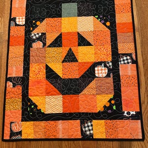 May include: A quilted wall hanging featuring a pumpkin design. The pumpkin is made of orange and yellow fabric squares, with a black background and border. The design includes a variety of patterns, including polka dots, stripes, and gingham. The quilt is machine-stitched.