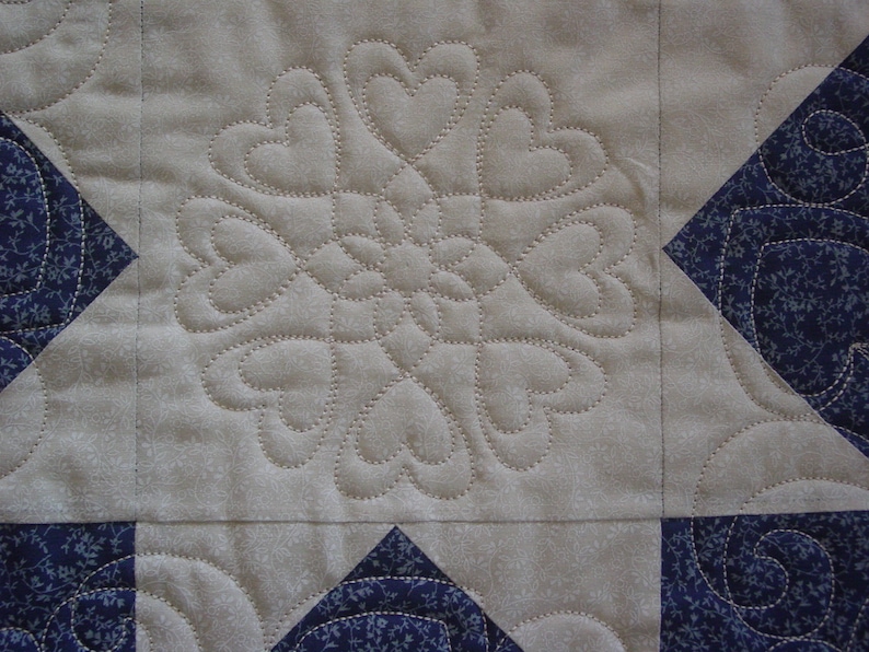 Puede incluir: Primer plano de una colcha con fondo blanco y estampado floral azul. La colcha est&aacute; acolchada con un dise&ntilde;o de coraz&oacute;n.