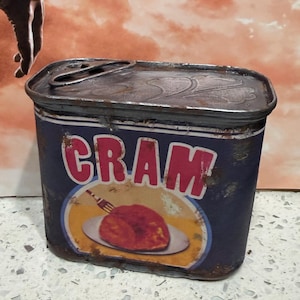 May include: A rectangular metal tin with a dark blue label featuring the word CRAM in large pink letters. Below the text is a graphic of a fork resting on a plate with a piece of food. The tin shows signs of rust and has a metal lid with a pull tab.