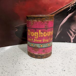 May include: A cylindrical metal can of Doghouse Beef Stew Dog Food with a weathered, rusted appearance. The label features a bright pink background with yellow text, a teal horizontal band, and a row of small yellow stars encircling the container.