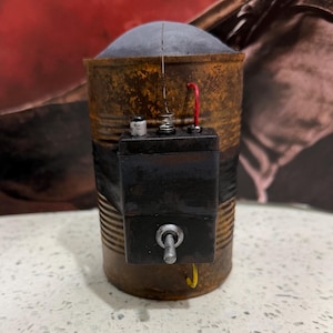 May include: A cylindrical, rusted metal container featuring a dark, rectangular electronic component attached to its side with black tape. The component includes a silver toggle switch, a small white button, a spring, and a red wire extending upward.