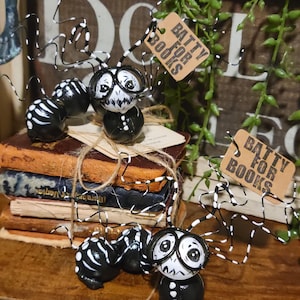 May include: Hand-painted black and white figurines with spider-like legs and faces, wearing glasses and a suit. The figurines are on a stack of old books tied with twine. Tags read "Batty for Books".