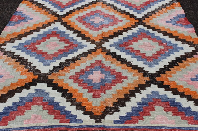 Pu&ograve; includere: Un tappeto colorato con un motivo geometrico in tonalit&agrave; di arancione, rosa, blu, marrone e bianco. Il tappeto ha un design a forma di diamante con un diamante centrale in ogni sezione.