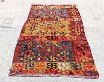 May include: A colorful, hand-woven rug with a geometric pattern in shades of red, orange, yellow, blue, and black. The rug has a fringed edge and is made of wool.