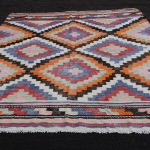 Pu&ograve; includere: Un tappeto vintage colorato con un motivo geometrico a rombi in tonalit&agrave; di arancione, rosa, blu e marrone. Il tappeto ha uno sfondo bianco e un bordo a frange.
