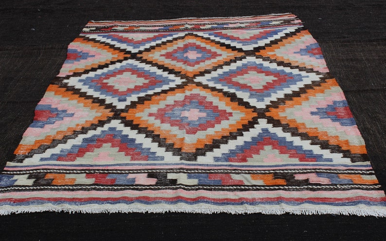 Pu&ograve; includere: Un tappeto vintage colorato con un motivo geometrico a rombi in tonalit&agrave; di arancione, rosa, blu e marrone. Il tappeto ha uno sfondo bianco e un bordo a frange.