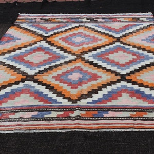 Pu&ograve; includere: Un tappeto turco vintage con un motivo geometrico in tonalit&agrave; di arancione, blu, rosa e marrone. Il tappeto ha uno sfondo bianco e un bordo a frange.