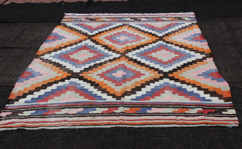 Pu&ograve; includere: Un tappeto turco vintage con un motivo geometrico in tonalit&agrave; di arancione, blu, rosa e marrone. Il tappeto ha uno sfondo bianco e un bordo a frange.