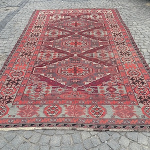 Puede incluir: Una alfombra tradicional de color rojo, gris y negro con diseños intrincados. La alfombra tiene un borde con flecos y está hecha de lana.