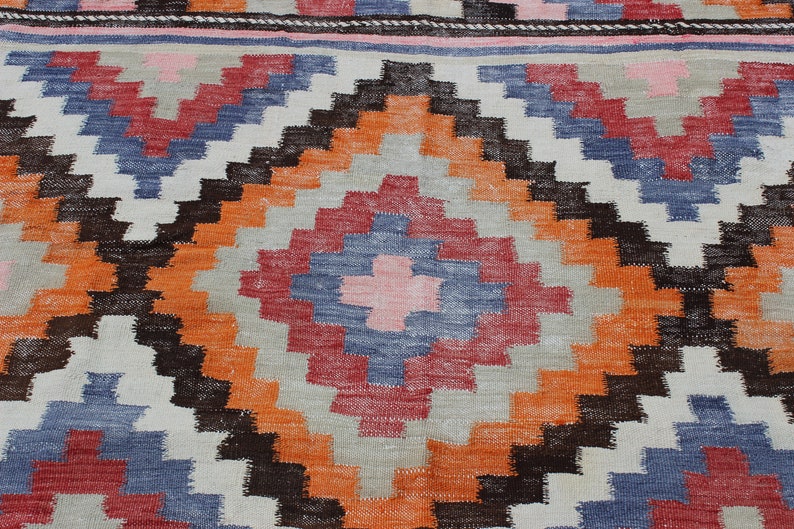 Pu&ograve; includere: Primo piano di un tappeto tessuto colorato con un motivo geometrico in tonalit&agrave; di arancione, rosso, blu, marrone e bianco.