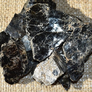 May include: A cluster of black mica mineral specimens with a reflective, layered appearance. The dark, shiny stones are arranged on a textured, tan burlap surface, creating a contrast in color and texture.