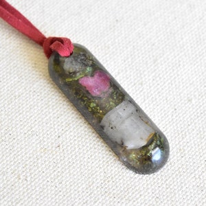 May include: A long, dark grey pendant with a red suede cord. The pendant is filled with clear resin and contains small pieces of pink, green, and white materials.