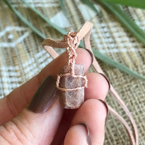 Könnte beinhalten: Ein roségoldener, drahtgewickelter Anhänger mit einem braunen und rosafarbenen Stein. Der Anhänger ist an einer braunen Lederschnur befestigt.