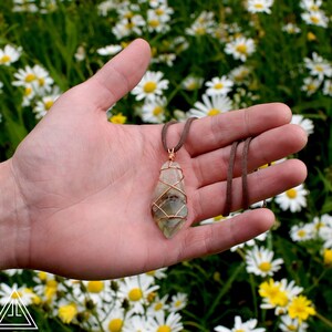 May include: A green and white crystal pendant wrapped in copper wire, hanging from a brown leather cord. The pendant is held in a hand against a background of white daisies.