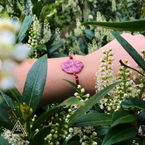 May include: A pink gemstone bracelet with a raw, irregular stone center, wrapped in gold-colored wire. The bracelet features a maroon cord and small red beads. The background is a lush green with white flowers.