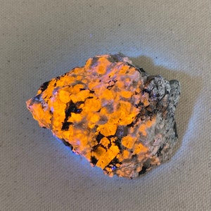 May include: A close-up of a rough, gray rock with a bright orange glow. The rock has a textured surface and appears to be a mineral specimen.
