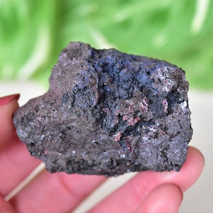Puede incluir: Una roca rugosa de color gris oscuro con matices iridiscentes azules y rojos. La roca tiene una superficie texturizada y parece ser una muestra mineral natural.