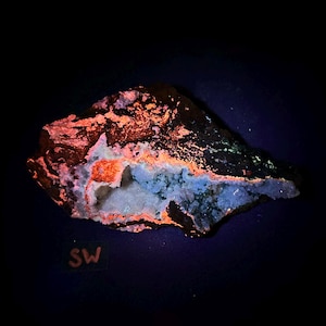 Op de afbeelding: Een close-up van een steen met een wit, kristallijn interieur. De steen gloeit oranje en groen onder UV-licht. De tekst "SW" staat op een zwart label in de linkerbenedenhoek van de afbeelding.